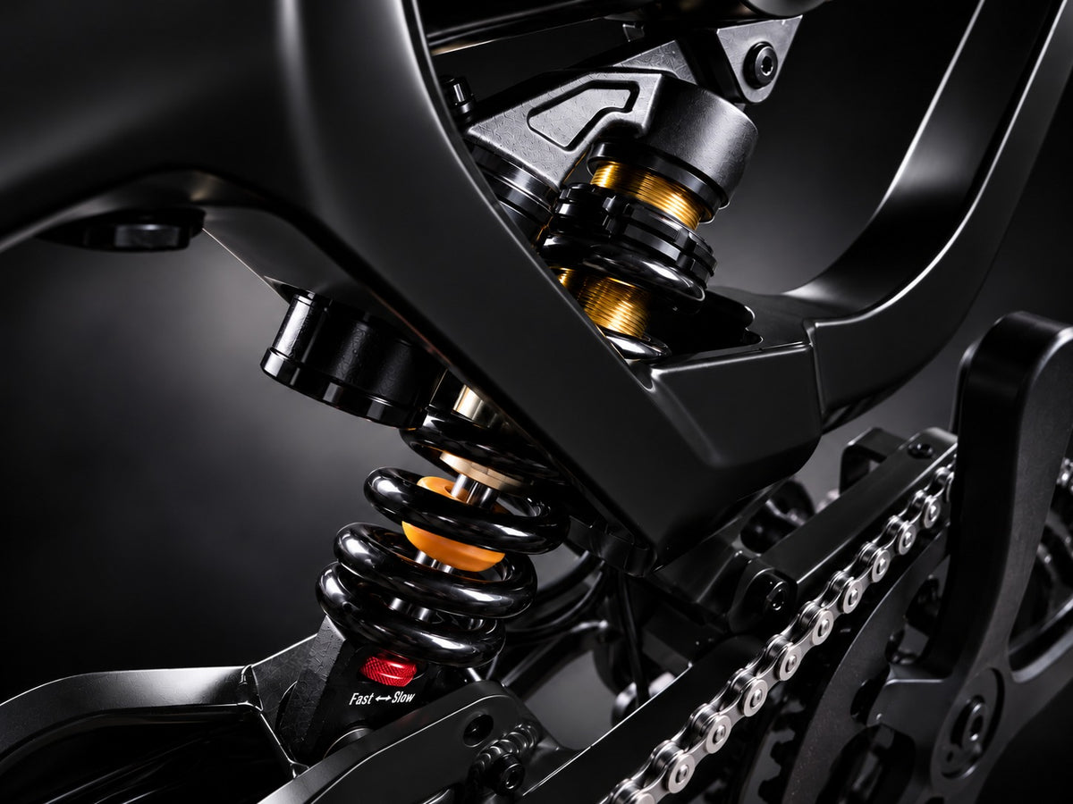 Close-up of a black motorcycle rear suspension system featuring a black coil spring with gold-colored shock absorber elements, set against a dark studio background.